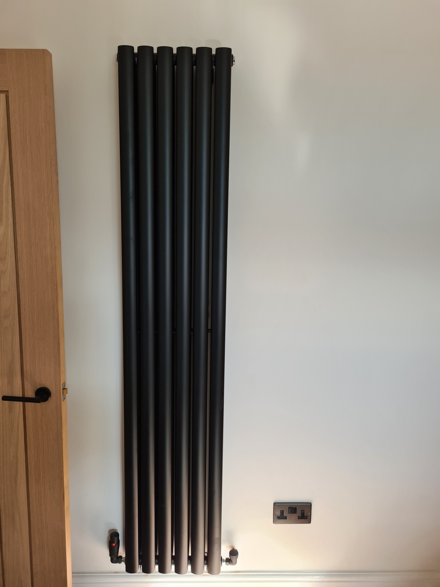 Black vertical column radiator installed in bedroom next to oak door
